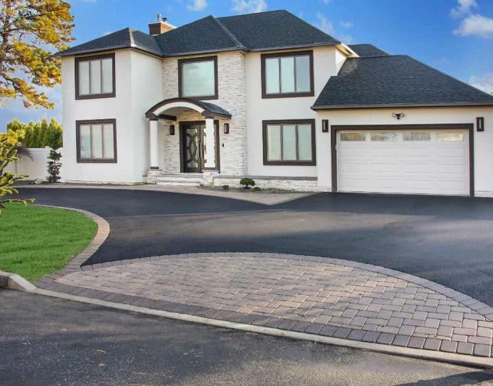 A modern two-story house with white walls, black trim, and a dark roof. The property features a large driveway and a garage, framed by a well-manicured lawn and trees.