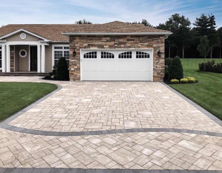 A house with a stone facade and a two-car garage, featuring a paved driveway bordered by grass and landscaping.
