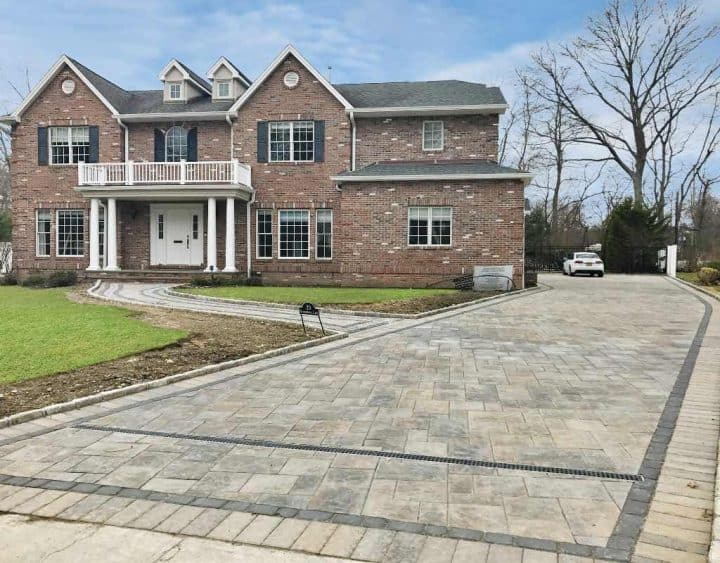 Large brick house with a white balcony and multiple windows. A long paved driveway leads to the garage. A car is parked near the trees in the background. Sparse landscaping in the yard.