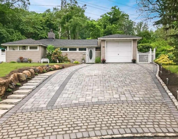 Single-story house with a gray roof and attached garage. Stone driveway with landscaped edges leads to the front. Trees and shrubs surround the property.