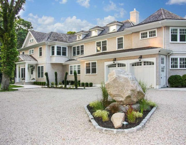Large two-story house with light-colored siding and a gravel driveway. There's a landscaped area with a large rock and plants in the foreground.