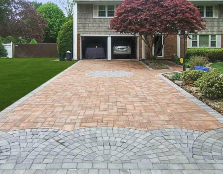 A brick driveway leads to a garage with a car parked halfway inside. A tree with red leaves stands to the right, and a green lawn is on the left.