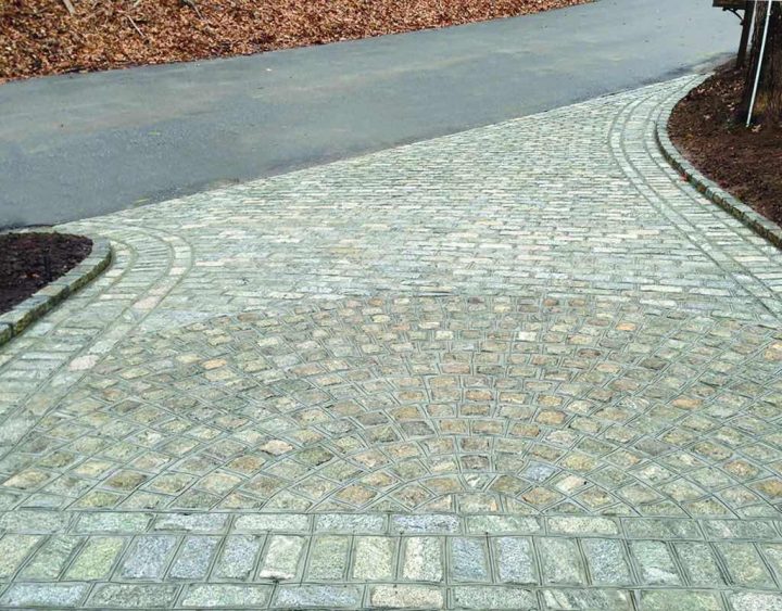 A stone-paved walkway with a circular pattern meets a paved road, surrounded by soil and fallen leaves.