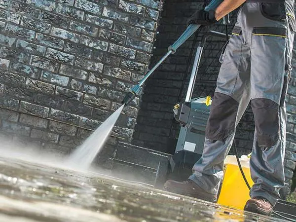 Person in workwear using a pressure washer to clean a wet stone or brick outdoor surface near a wall.