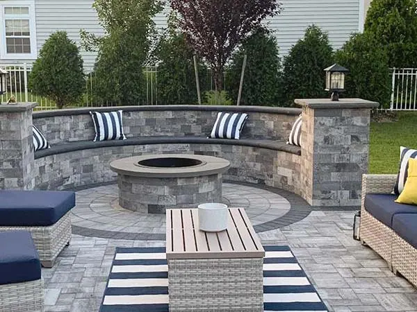Outdoor patio with a curved stone bench, striped cushions, a circular fire pit, a coffee table, and wicker seating on a paved surface with a striped rug.