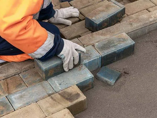 A person wearing gloves and an orange jacket is laying rectangular bricks or pavers in a sand bed.