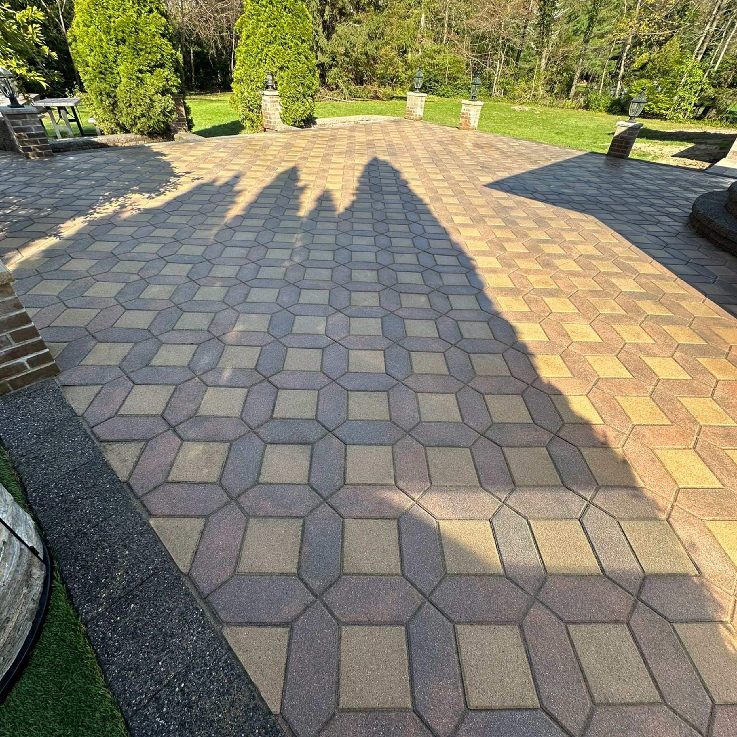 A geometric-patterned patio with rectangular and hexagonal pavers, casting sharp, tall shadows from nearby trees on a sunny day.