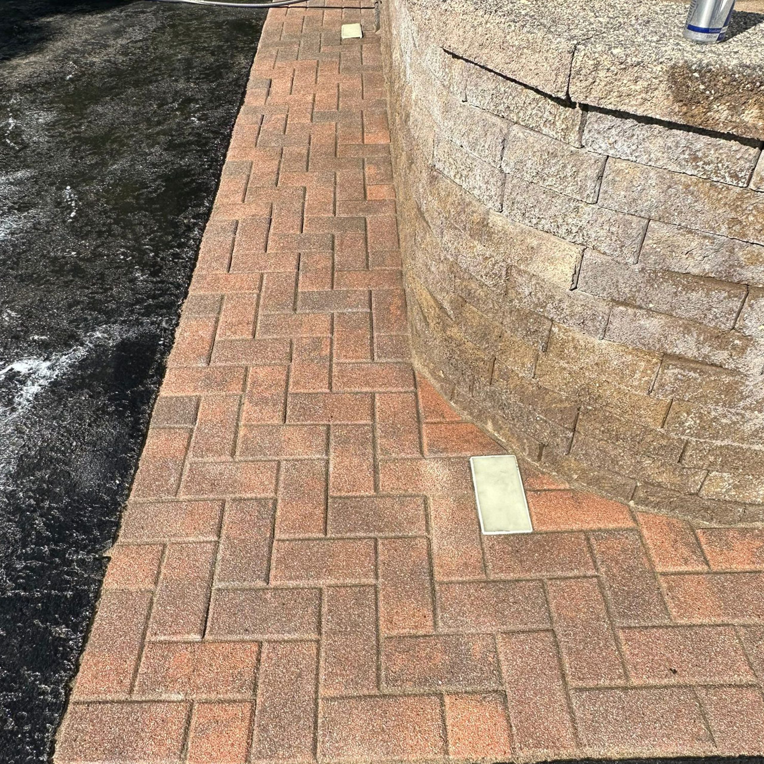 Brick walkway with a herringbone pattern runs alongside a curved stone wall, with two rectangular light fixtures embedded in the walkway.