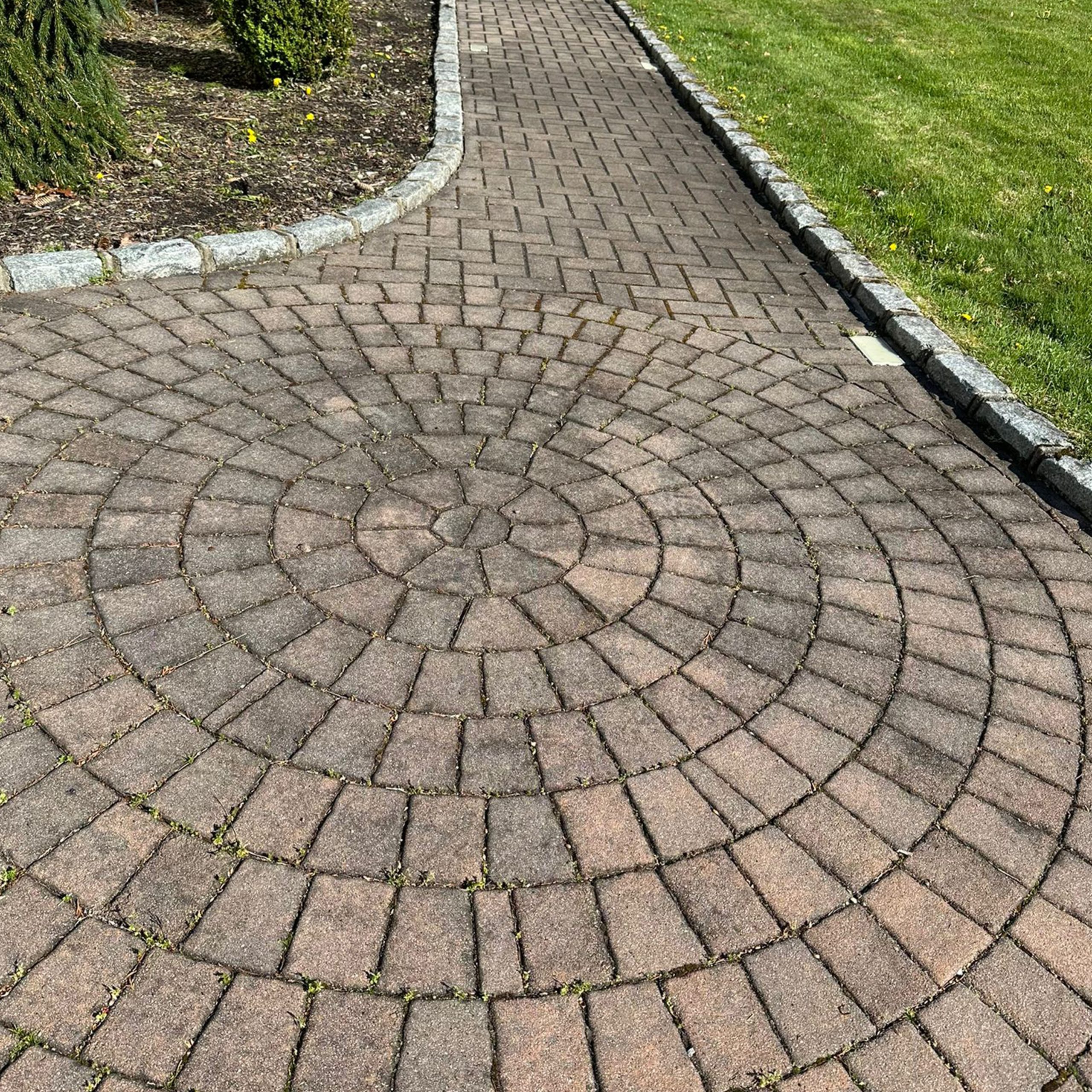 A brick walkway with a circular pattern in the foreground, bordered by grass and a garden bed with mulch and plants.