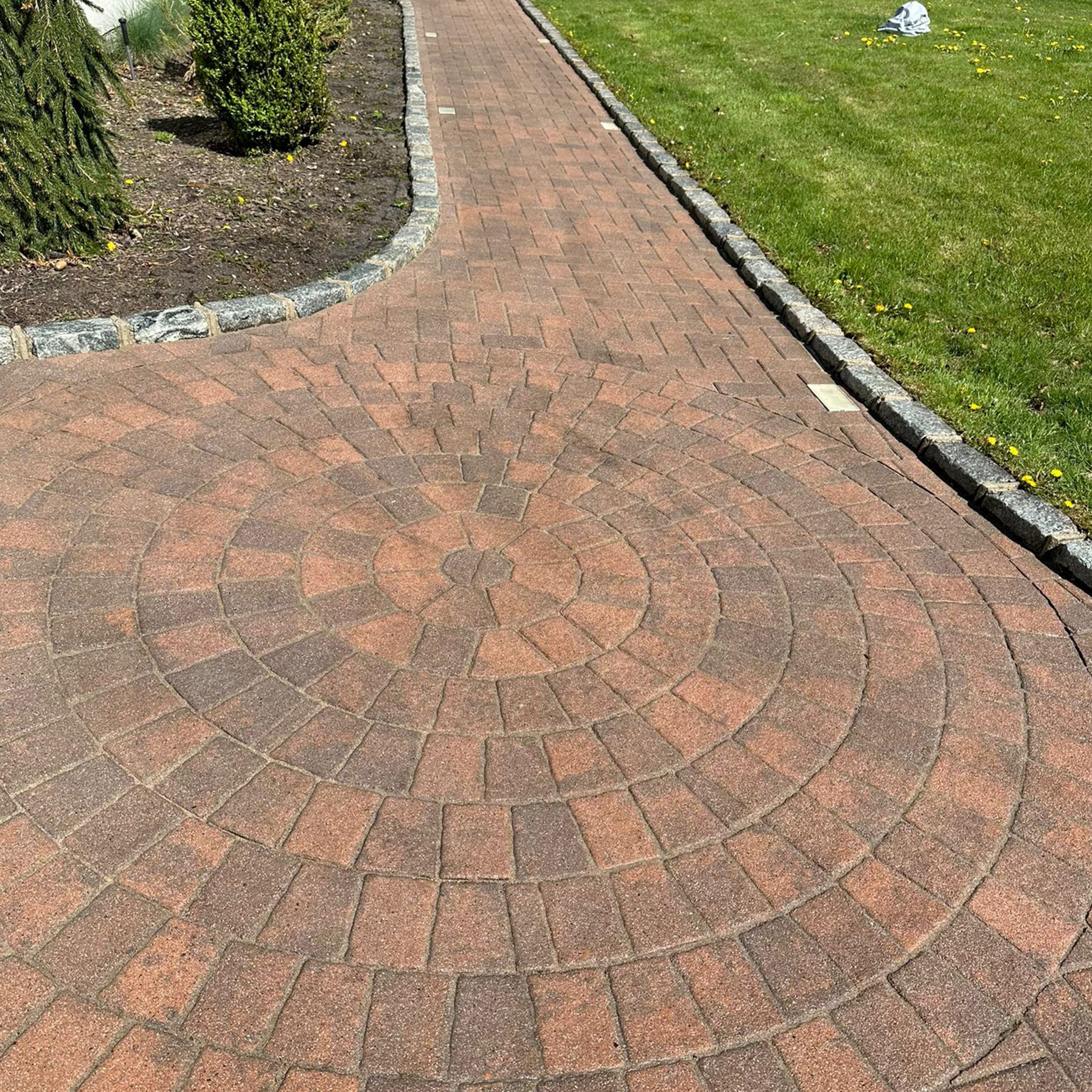 Brick walkway with a circular pattern in the center, bordered by a lawn on the right and a garden bed with mulch and plants on the left.