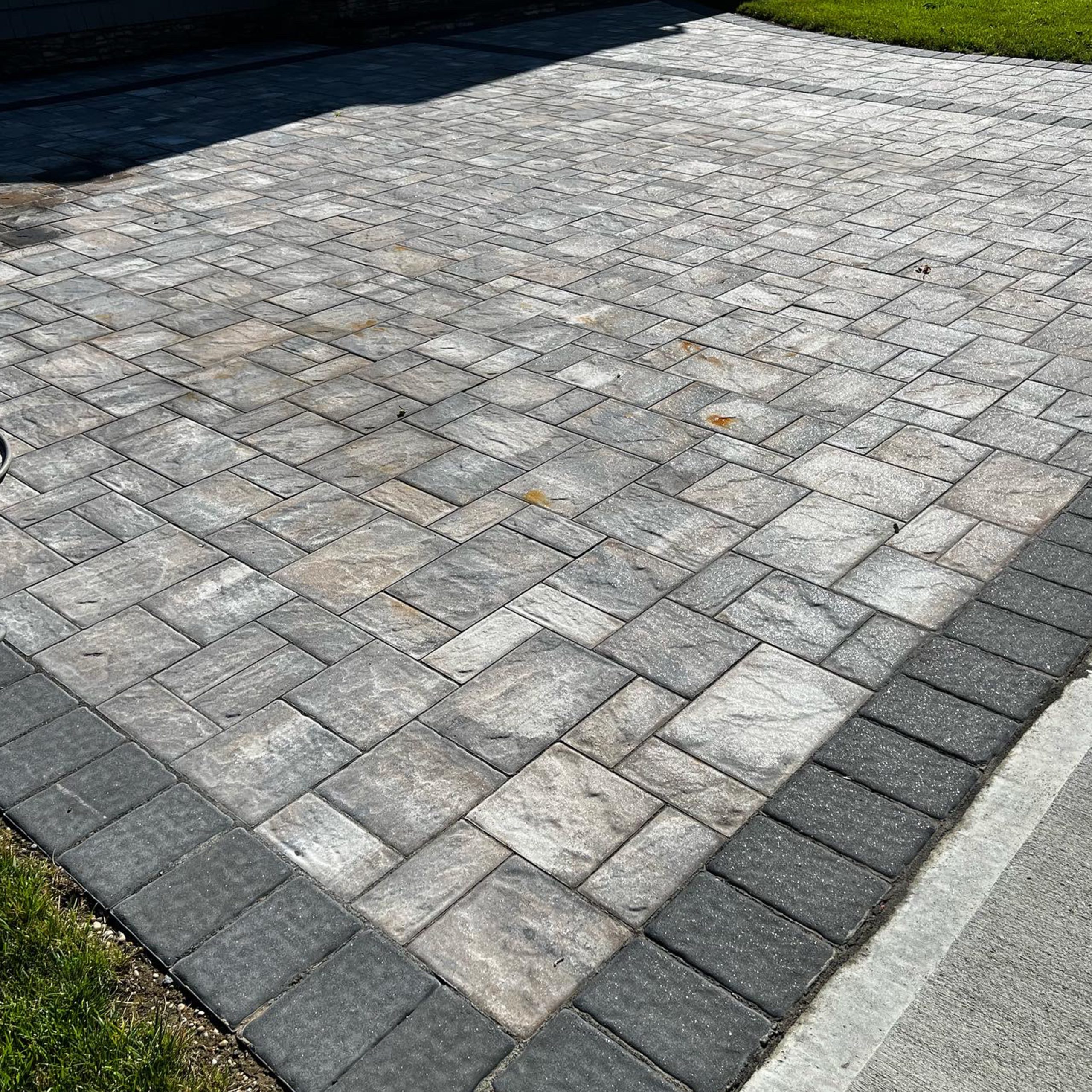 Rectangular stone pavers arranged in a geometric pattern form a driveway bordered with darker stones; grass and concrete edge are visible.